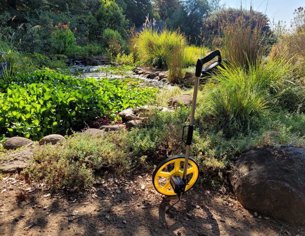 A landscape measuring wheel in front of a pond and garden