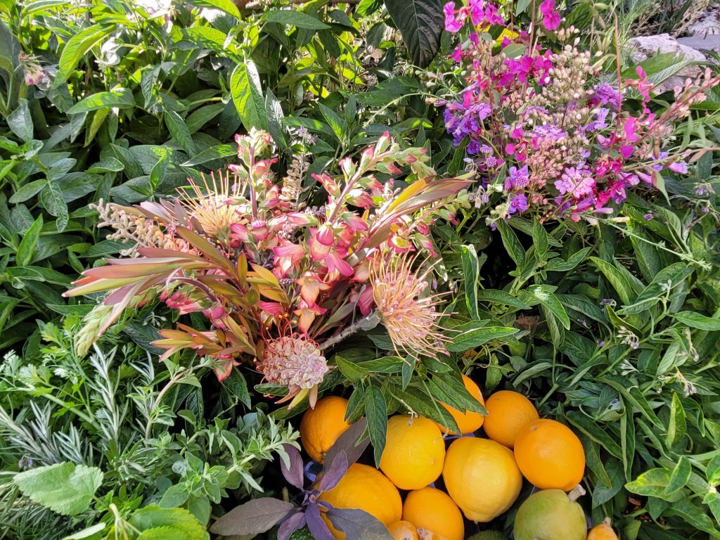2 colorful flower arrangements sit outside on green foliage next to a platter of fruit